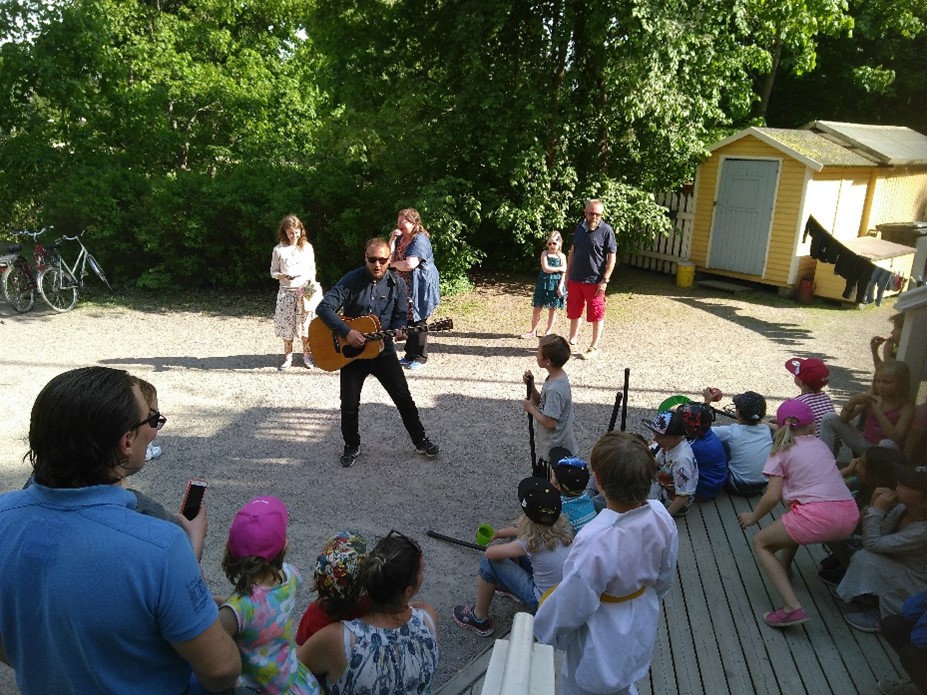 mannen-spelar-gitarr-pa-garden-framfor-en-grupp-barn-och-vuxna